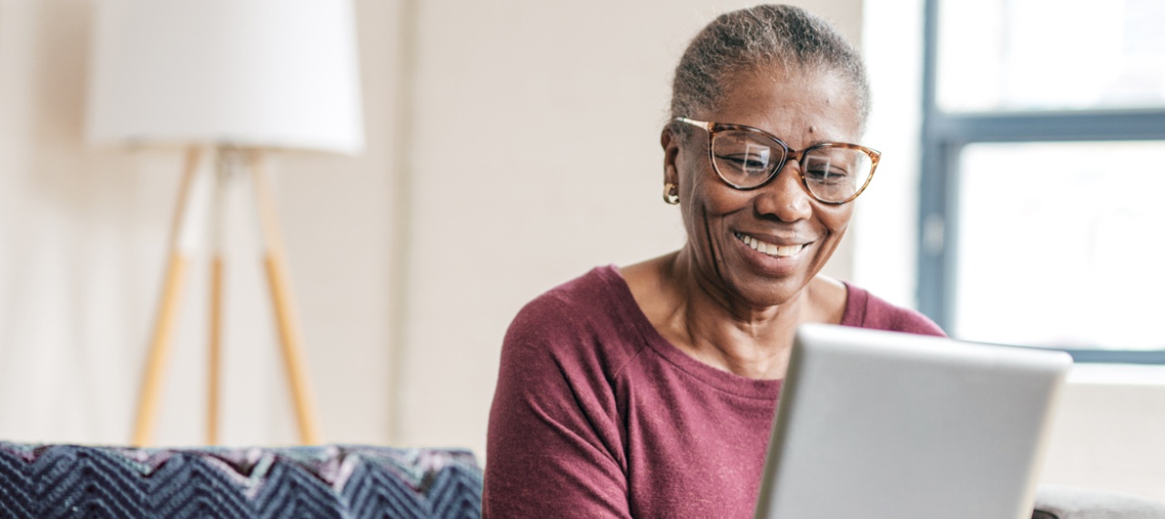 A smiling older woman with short gray hair and glasses sits comfortably on a couch, looking down at a tablet in her hands. Soft natural light and a calm home setting create a warm, approachable moment of focused engagement with technology.