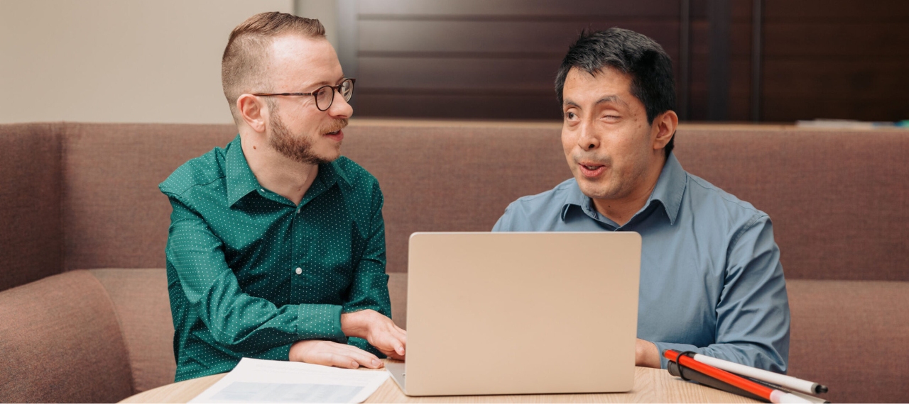 Two native user testers review digital assets on a laptop. One’s white cane rests on the table as they discuss, illustrating how Onward Accessibility engages professionals with disabilities for testing in its digital accessibility solutions case study.
