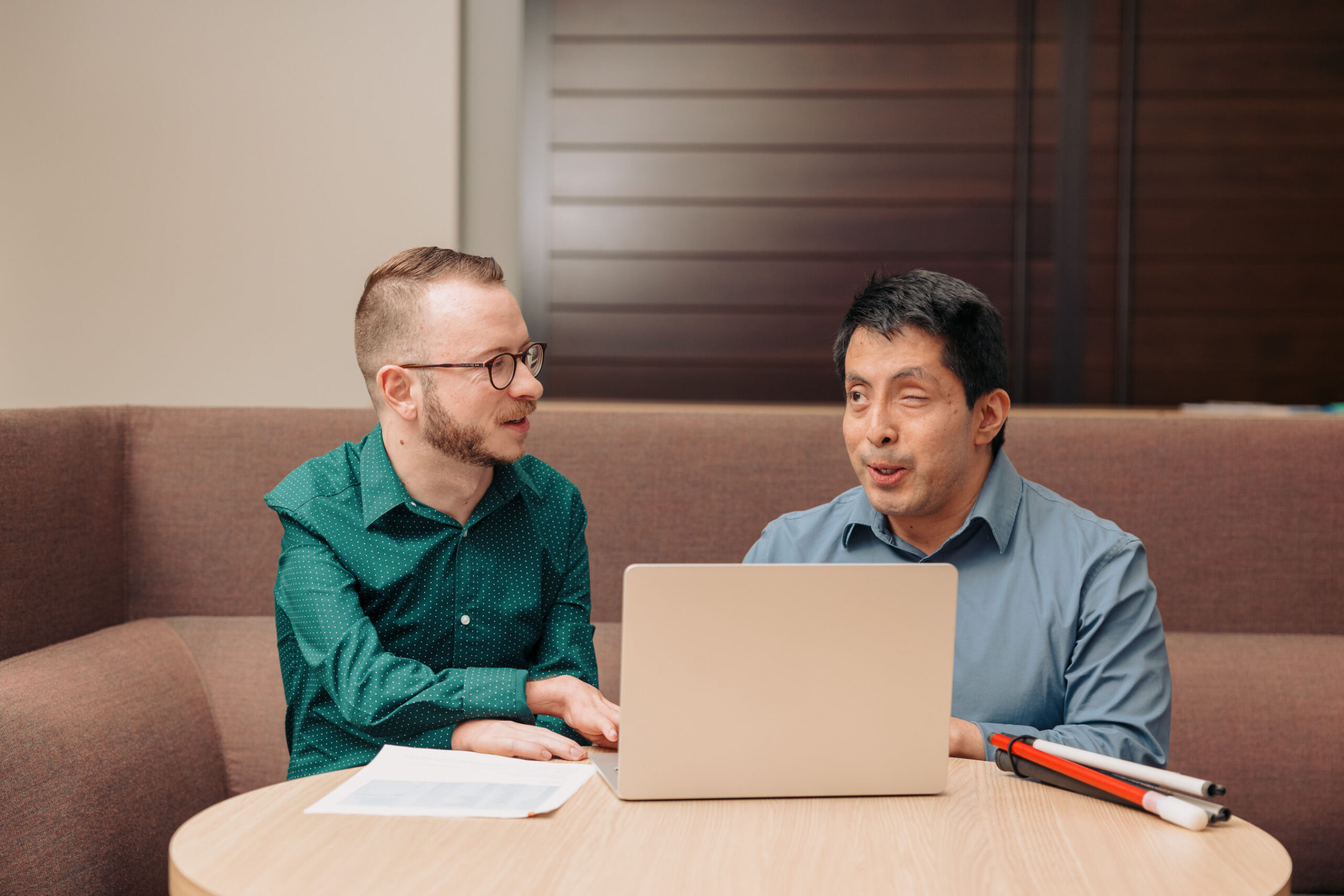 Two native user testers review digital assets on a laptop. One’s white cane rests on the table as they discuss, illustrating how Onward Accessibility engages professionals with disabilities for testing in its digital accessibility solutions case study.