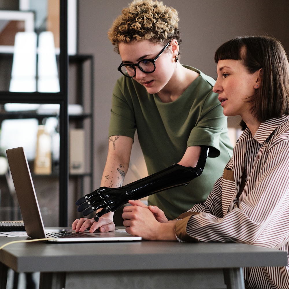 Young woman with prosthetic arm pointing at laptop and talking to her colleague during work at office