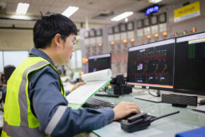 In an energy control room, a professional in a high-visibility safety vest reviews live grid data across dual monitors while referencing handwritten notes at his workstation. This case study image demonstrates the risk of relying on manual workarounds and memory under pressure, highlighting the need to embed critical information directly within accessible software.