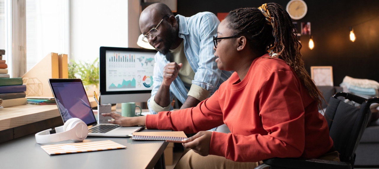 Two colleagues collaborate at a desk, reviewing data on a laptop while a nearby monitor displays charts and analytics dashboards. The scene reflects a focused, team-oriented approach to problem-solving in a modern, well-lit workspace.