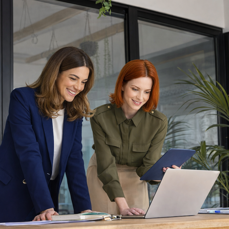 Two professionals stand at a wooden table in a modern office, reviewing a laptop and documents. The person on the left wears a navy blue blazer, while the person on the right wears an olive-green top and holds a tablet. Large glass walls and indoor plants are visible in the background.